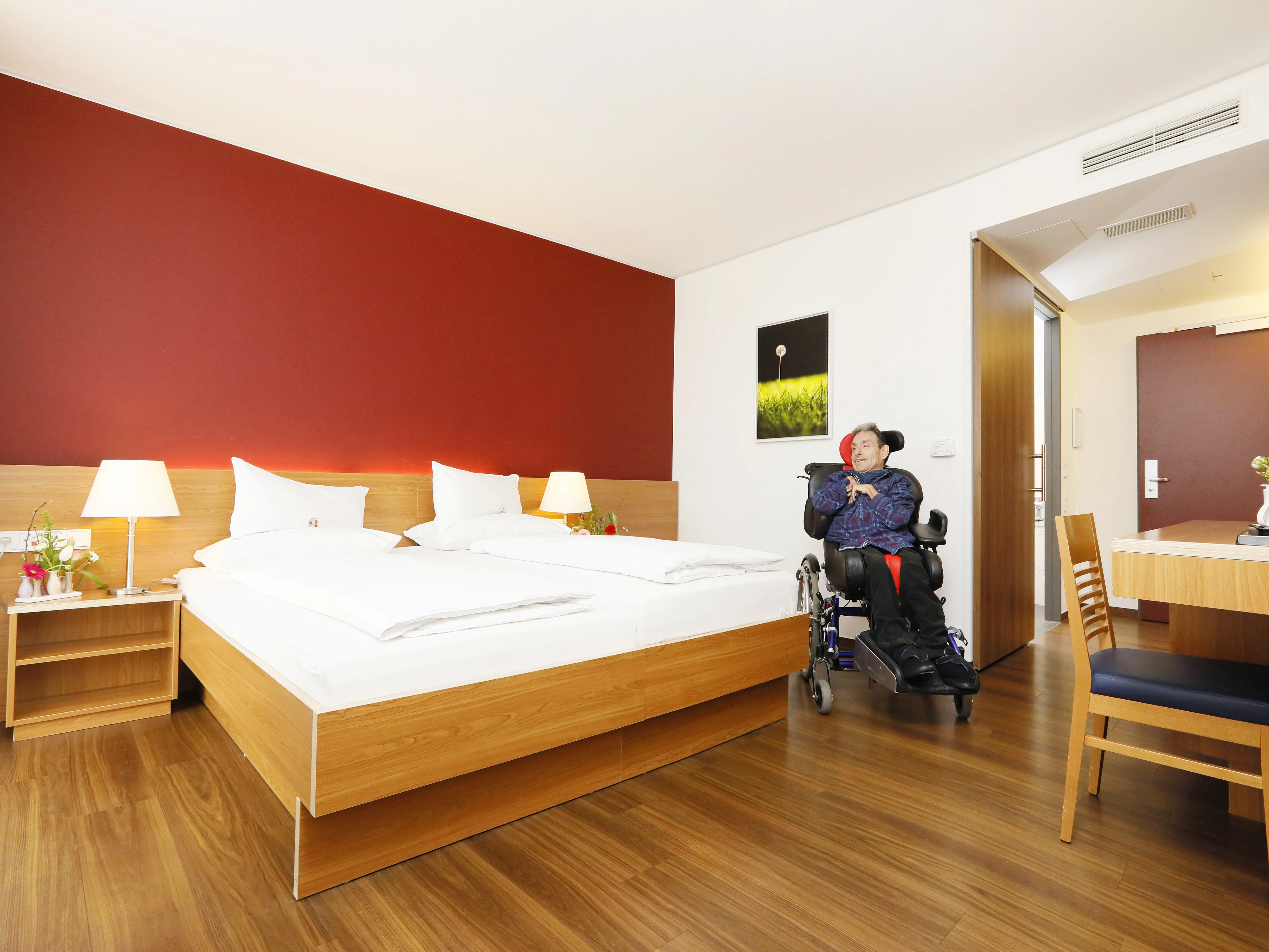 Barrierefreies Hotelzimmer mit roter Akzentwand, Doppelbett, Rollstuhlfahrer, Holzboden.