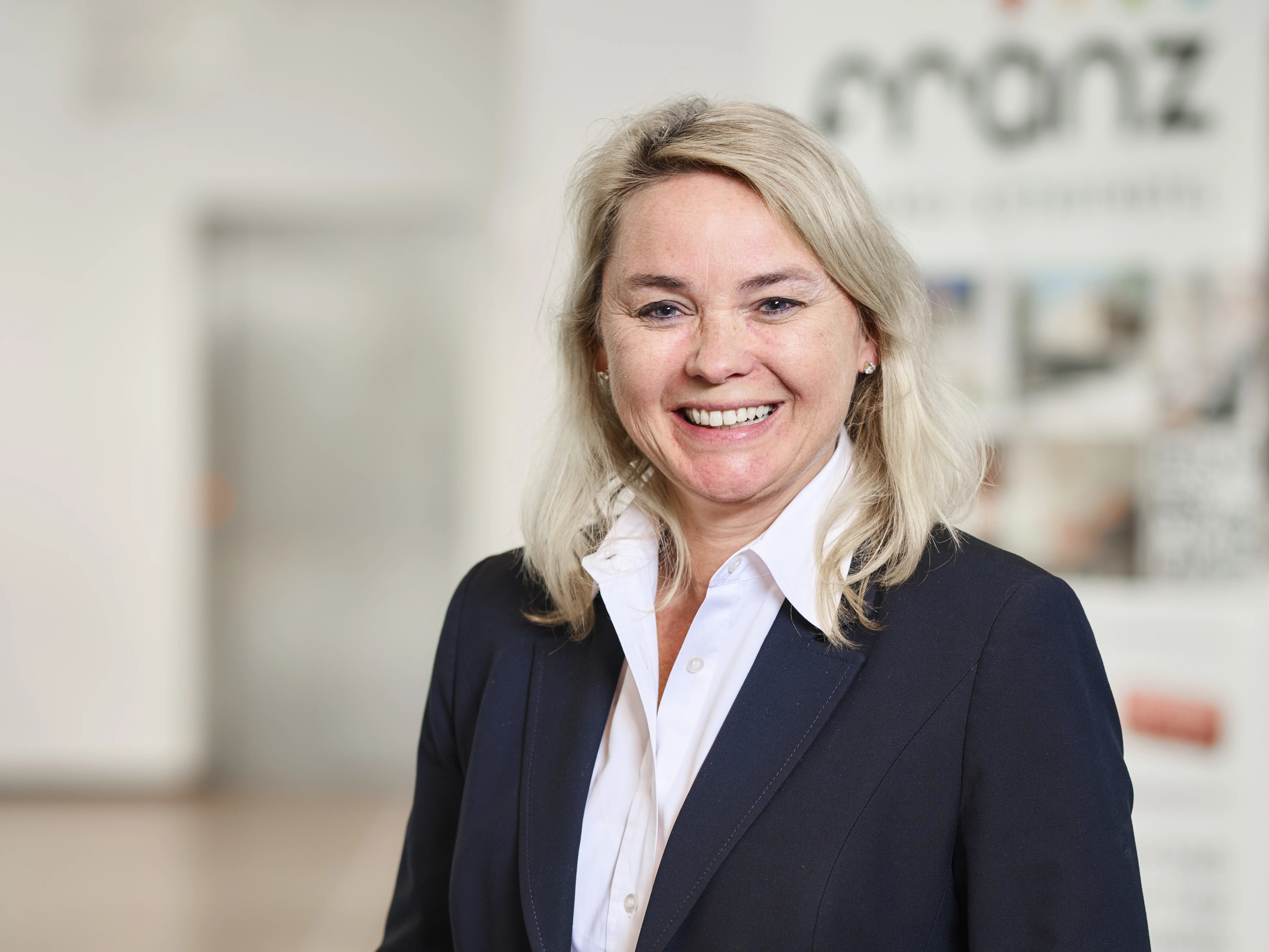 Lächelnde Frau im Business-Outfit vor verschwommenem Hintergrund.