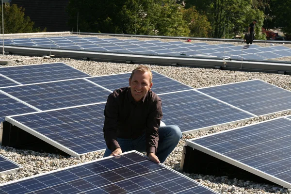 Mann kniet zwischen Solarpaneelen auf einem Kiesdach, sonniger Tag.