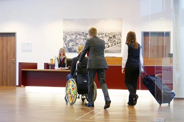 Person am Empfangstresen, zwei Personen mit Gepäck, eine davon im Rollstuhl.